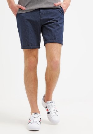 Uomo con pantaloncini blu navy a pois, maglietta grigia e sneakers bianche Adidas con strisce nere e rosse, in piedi con le mani in tasca.