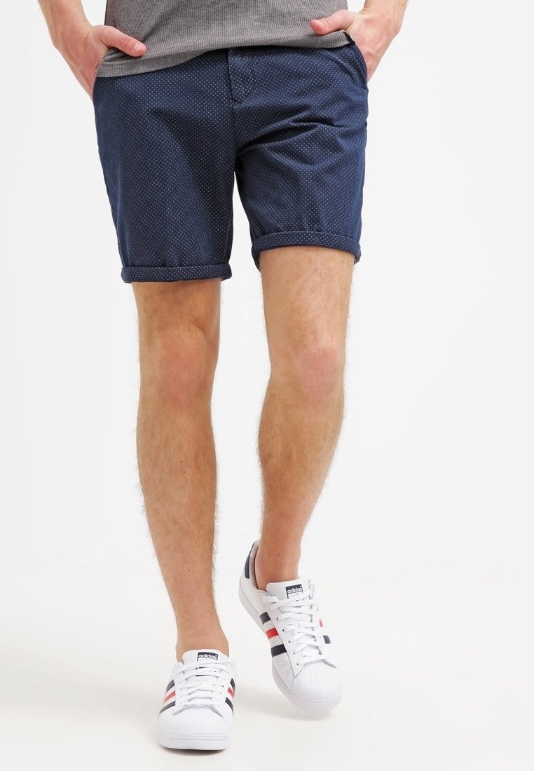 Homme portant un short bleu marine à pois, un haut gris et des baskets blanches Adidas avec des bandes noires et rouges, debout les mains dans les poches.