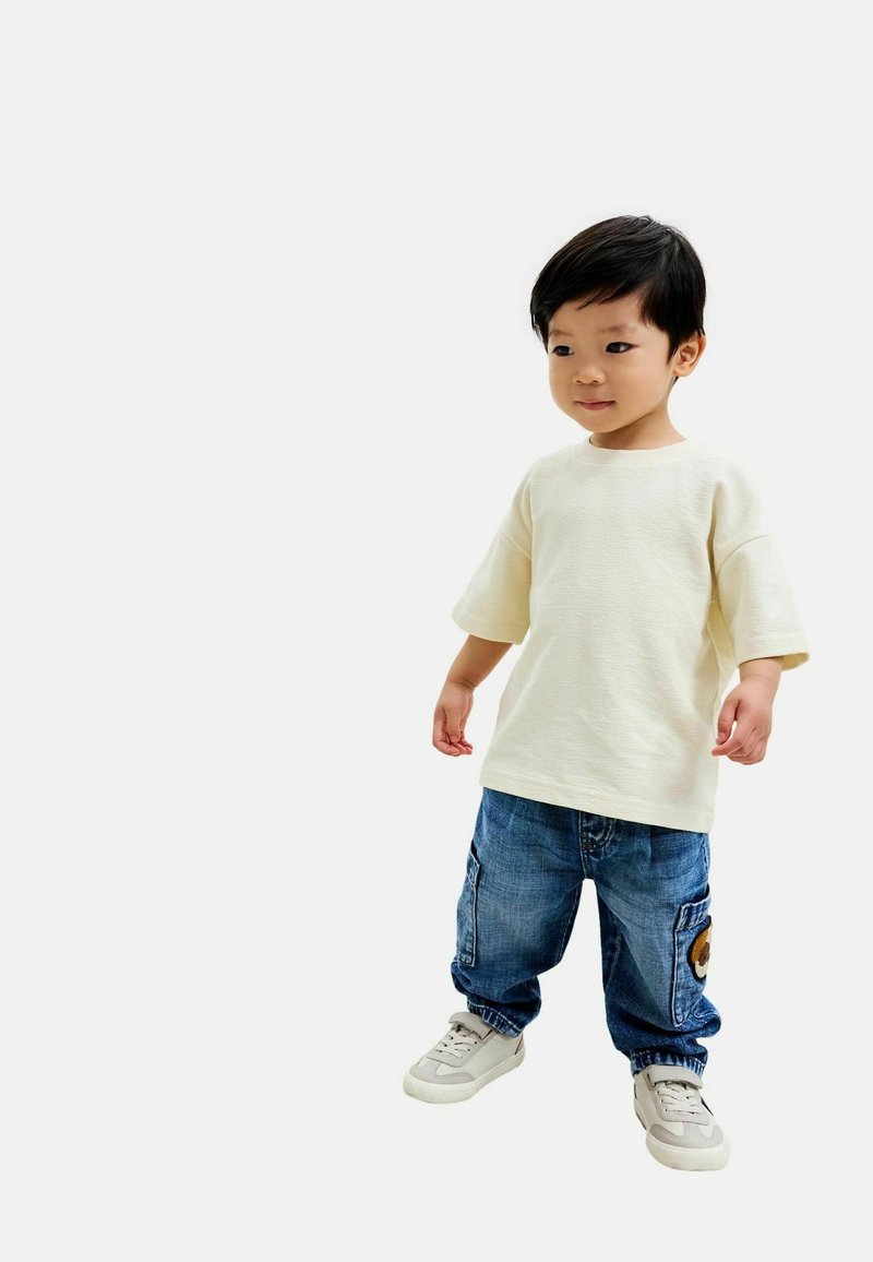 Top corto a maniche corte color crema chiaro, joggers in denim blu con tasche e sneakers bianche con accenti grigi. L'outfit ha un design casual.