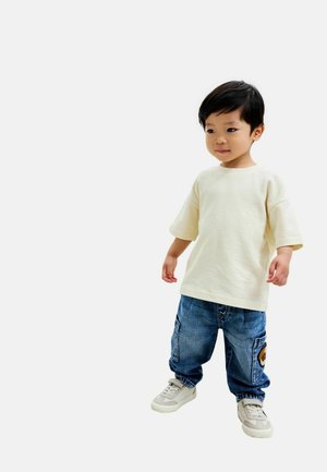 Junges Kind steht, trägt ein hellbeiges Hemd, blaue Cargo-Jeans mit einem Flicken und weiße Sneaker auf neutralem Hintergrund.