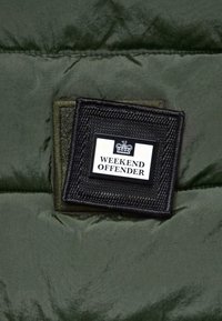 Vihreä puhallettava takki, jossa on teksturoitu musta läiskä, jossa lukee "WEEKEND OFFENDER" ja kruunulogo. Liukas kangas, jossa on hieman kiiltoa.