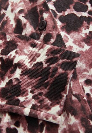 Hedvábná košile s ribovanou strukturou v růžové, krémové a černé leopardí potisku; má černý knoflíkový detail na přední straně a texturované rukávy.