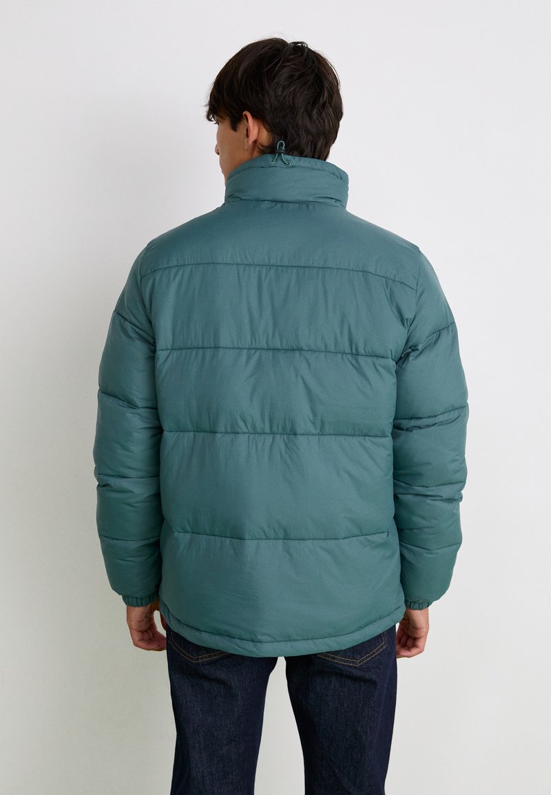 Grüne Puffjacke mit hohem Kragen, gestepptem Design, elastischen Bündchen und leicht lockerer Passform, getragen über dunklen Jeans.