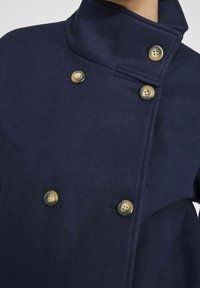 Marineblå ull peacoat med høy krage, med dobbelrad med messingknapper og en myk tekstur.