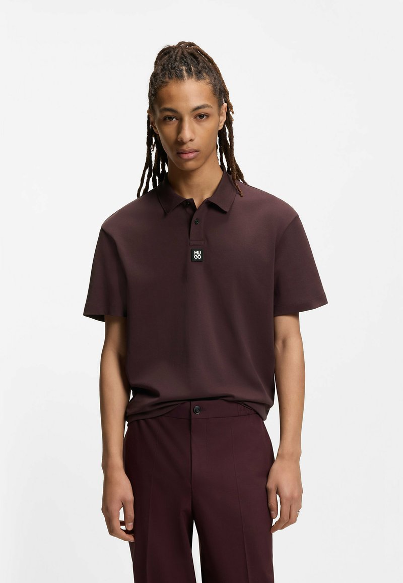 Jeune homme aux dreadlocks longs portant un polo marron foncé et un pantalon assorti, debout devant un fond blanc uni.