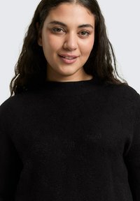 Jonge vrouw met lang donker haar lacht, draagt een zwarte trui met een subtiel getextureerd luipaardpatroon op een lichtgrijze achtergrond.