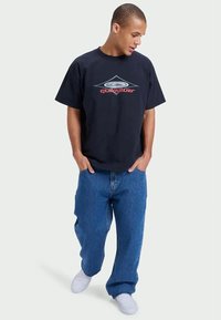 Melna kokvilnas T-kreklu ar grafisku dizainu, kurā attēlots dimanta formas elements un zīmola nosaukums "Quiksilver" sarkanā krāsā. Apvienots ar ziliem džinsu biksēm.