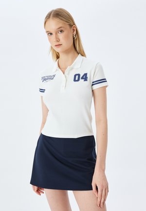 Valkoinen lyhythihainen polo-paita, jossa tummansiniset raidat hihoissa ja printti "04"; yhdistettynä tummansiniseen mini-hameeseen, jossa on nappikoristeita.