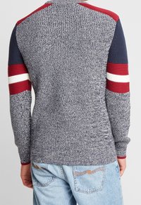 Pull en tricot bleu marine et gris avec des accents rayés rouges et blancs sur les manches. Dispose de poignets côtelés et d'un tissu texturé.