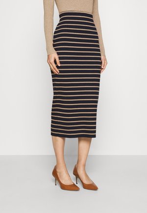 Kobieta w czarnej spódnicy za kolano z beżowymi poziomymi paskami, beżowej bluzce z długim rękawem i brązowych wysokich szpilkach ze spiczastymi noskami stojąca na szarej podłodze.