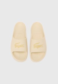 Beige slip-on sandaler med texturerad sula och en guldfärgad krokodil-logotyp på remmen. Slät, gummi-liknande material hela vägen.