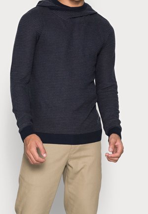 Homme portant un pull côtelé bleu marine foncé ajusté avec capuche et un pantalon beige sur un fond clair uni.