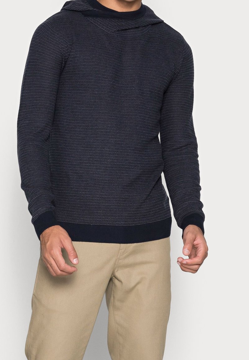 Homme portant un pull côtelé bleu marine foncé ajusté avec capuche et un pantalon beige sur un fond clair uni.