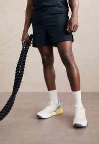 Scarpe atletiche bianche e grigie con accenti gialli, abbinate a pantaloni corti neri a trama e calze bianche, mentre si tiene una corda da esercizio nera.