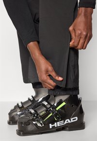 Schwarze Skischuhe mit grünen Akzenten und metallischen Schnallen, neben schwarzen Hosen mit elastischen Bündchen, die am Knöchel angepasst werden.