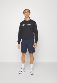 Sudadera negra con logo "Champion", pantalones cortos azul marino, calcetines blancos y zapatillas deportivas. Diseño simple con colores de logo en contraste.