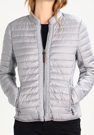 Donna che indossa una giacca trapuntata grigio chiaro con cerniera frontale e mani nelle tasche con zip, abbinata a pantaloni neri.