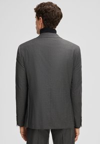 Mann in einem dunkelgrauen Nadelstreifenanzug und schwarzem Rollkragenpullover, der mit dem Rücken zu einem einfarbigen hellgrauen Hintergrund steht.