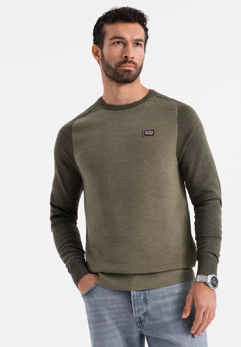 Maglione verde con texture a coste, collo rotondo e maniche a contrasto. Presenta una patch del logo sul petto e polsini aderenti. Indossato con jeans chiari.