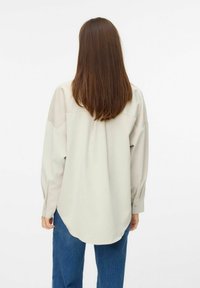 Light beige oversized shirt with a rounded hem and long sleeves, featuring shoulder seams and a smooth, soft texture. Worn with blue jeans.
