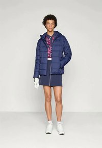 Marineblå pufferjakke med hette, med quiltet design, kombinert med en rosa blomsteroverdel og et marineblått skjørt. Modellen har på seg hvite sneakers og hansker.