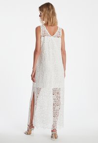 Langes weißes Spitzenkleid mit V-Ausschnitt, verziert mit Blumenmustern, kniehohen Schlitzen an beiden Seiten und einer halbtransparenten Textur.