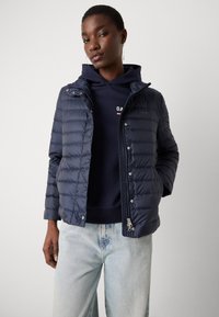Marineblå dunjakke med quiltet design, lynlås og trykknapper. Har hætte og er kombineret med en marineblå hættetrøje og lyse jeans.