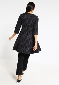 Mujer caminando alejándose, vestida con un abrigo acolchado negro de mangas tres cuartos, pantalones cortos negros y zapatos negros sin cordones con suelas blancas.