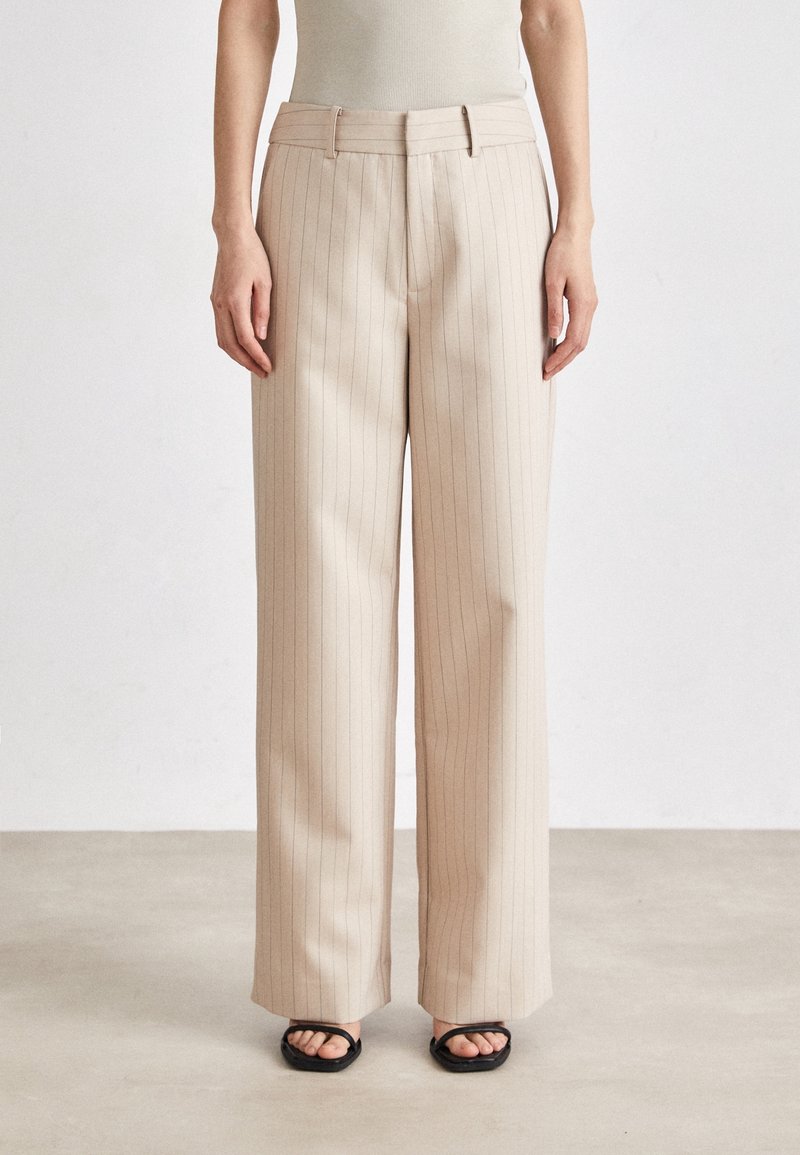 Femme portant un pantalon large beige à rayures fines et des sandales noires à bout carré, debout devant un fond clair uni.