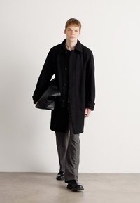 Cappotto lungo nero, con frontale bottonato, dal tessuto liscio, indossato sopra un top scuro e pantaloni a righe grigie. Scarpe nere, in possesso di una borsa nera.