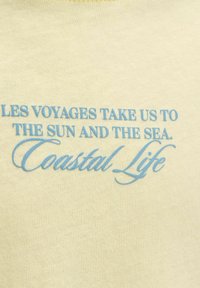 Μπλε κείμενο σε μπεζ ύφασμα γράφει: «Les voyages μας ταξιδεύουν στον ήλιο και τη θάλασσα. Παραθαλάσσια ζωή» με ανάμεικτες γραμματοσειρές.