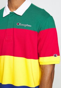 Polo shirt coloré avec des rayures horizontales en vert, rouge, jaune et bleu. Fabriqué en coton avec un col blanc et un logo brodé.