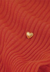 Zacht geribbeld stof in levendig rood met een kleine gouden hartvormige accentspeld. De textuur is glad met delicate ribbels.