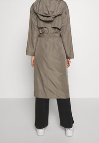 Cappotto lungo khaki con cappuccio, vita cintrata e maniche lunghe. Presenta una spalla posteriore e un tessuto liscio e resistente all'acqua. Indossato sopra pantaloni neri.