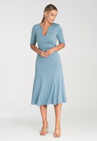 Helles blaues Kleid mit V-Ausschnitt und kurzen Ärmeln. Ausgestellter Rock, glattes Material und tailliertes Oberteil. Kombiniert mit klaren Blockabsatzsandalen.