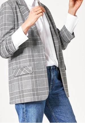 Person iført en grå ternet blazer, hvid skjorte med knapper og blå jeans, med hænder hævet i brysthøjde.