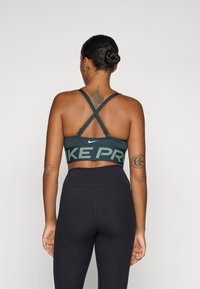 Nike Pro športni modrček v temno zeleni barvi s križnimi naramnicami, ki ima velik napis "NIKE PRO" v svetlo zeleni barvi. Gladek, raztegljiv material.