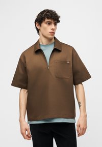 Νεαρός με σγουρά μαλλιά, φοράει καφέ κοντομάνικο πουκάμισο με φερμουάρ πάνω από ανοιχτό μπλε μπλουζάκι, μαύρο παντελόνι και στέκεται μπροστά σε απλό φόντο.