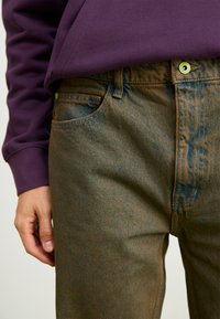 Denim jeans in faded blue with a textured finish, featuring green accents on hardware and a relaxed fit, paired with a purple sweatshirt.