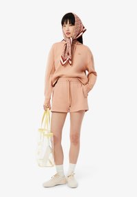 Pulover piersică și pantaloni scurți dintr-un material moale, asortați cu o eșarfă cu model și o geantă transparentă cu accente galbene. Adidași și șosete albe.