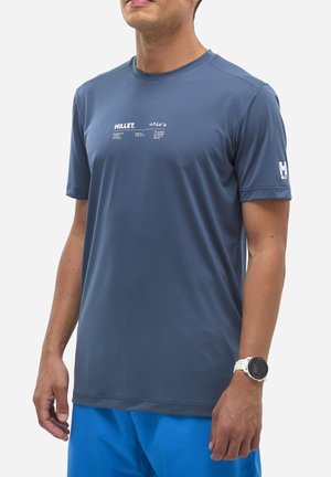 Homme portant un t-shirt de sport Millet bleu à manches courtes et un short bleu, avec une montre connectée blanche au poignet gauche.