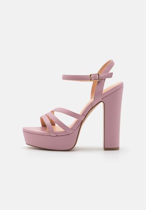 Lilla høje hæle plateau sandal med ankelrem, spænde lukning og tre slanke forremme på hvid baggrund.