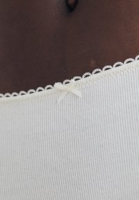 Țesătură ribbed de culoare gri deschis, cu un dantelă albă delicată pe margini și un accent de fundiță mică la talie, evidențiind textura și detaliile.