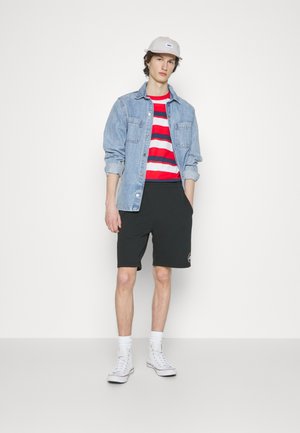 Chaqueta de mezclilla sobre una camiseta de rayas rojas y blancas, pantalones cortos negros con logo y zapatillas altas blancas. Fondo neutro.