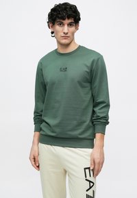 Zöld pulóver hosszú ujjal, kerek nyakkivágással és diszkrét EA7 logóval a mellrészen. Világos színű nadrággal párosítva, amelyen feltűnő logó található.