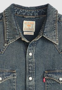 Cămașă din denim cu guler ascuțit, spălare albastră, două buzunare pe piept, butoane albastre tip snap și o etichetă roșie Levi's pe buzunarul din stânga.