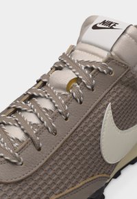 Κοντινό πλάνο μπεζ αθλητικού παπουτσιού Nike με υφή από δίχτυ, χοντρά δίχρωμα κορδόνια, λευκό σήμα swoosh και ταμπελάκι με το λογότυπο στη γλώσσα.