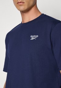 Marineblå bomulds T-shirt med et hvidt Reebok-logo på venstre bryst, rund hals og korte ærmer. Glat tekstur.