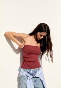 Top strapless burgundy cu plisuri laterale, asociat cu blugi din denim deschis și o cămașă legată în jurul taliei, albă cu dungi albastre.