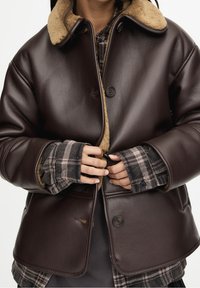 Veste en cuir marron avec un col en fourrure, dotée de deux boutons et d'un design matelassé, superposée à une chemise à carreaux.